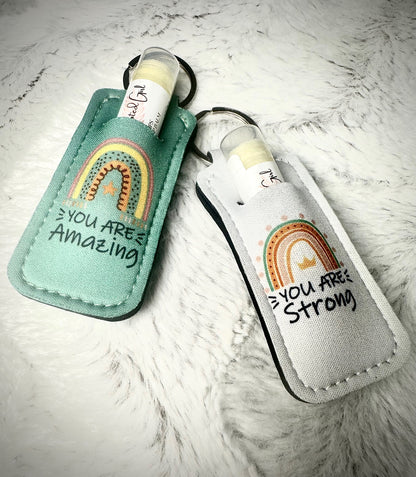 Two motivational keychains/lip balm holders with rainbow designs and text on a textured surface